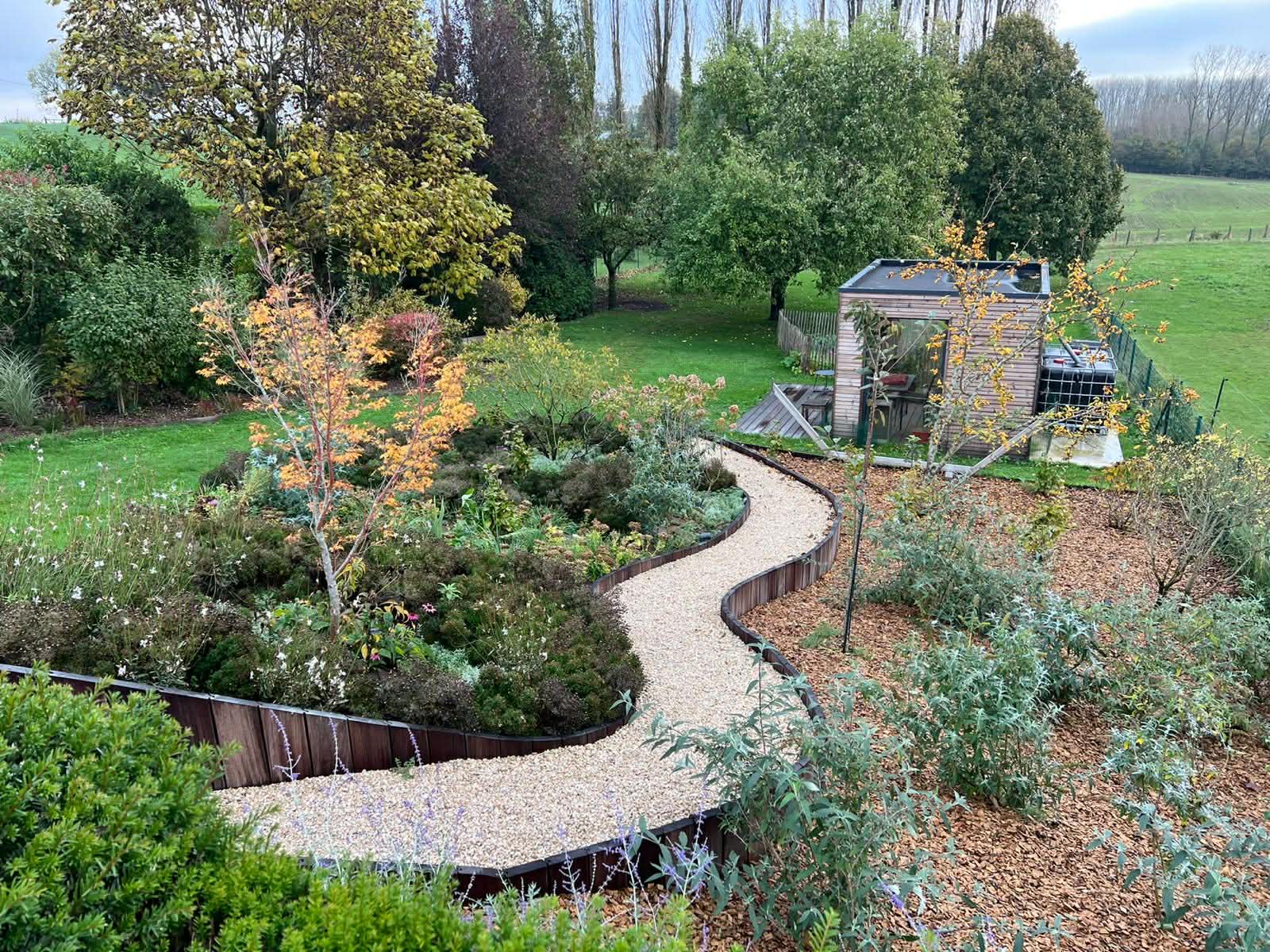 Jardin paysager avec allée sinueuse en gravier, massifs plantés et abri de jardin